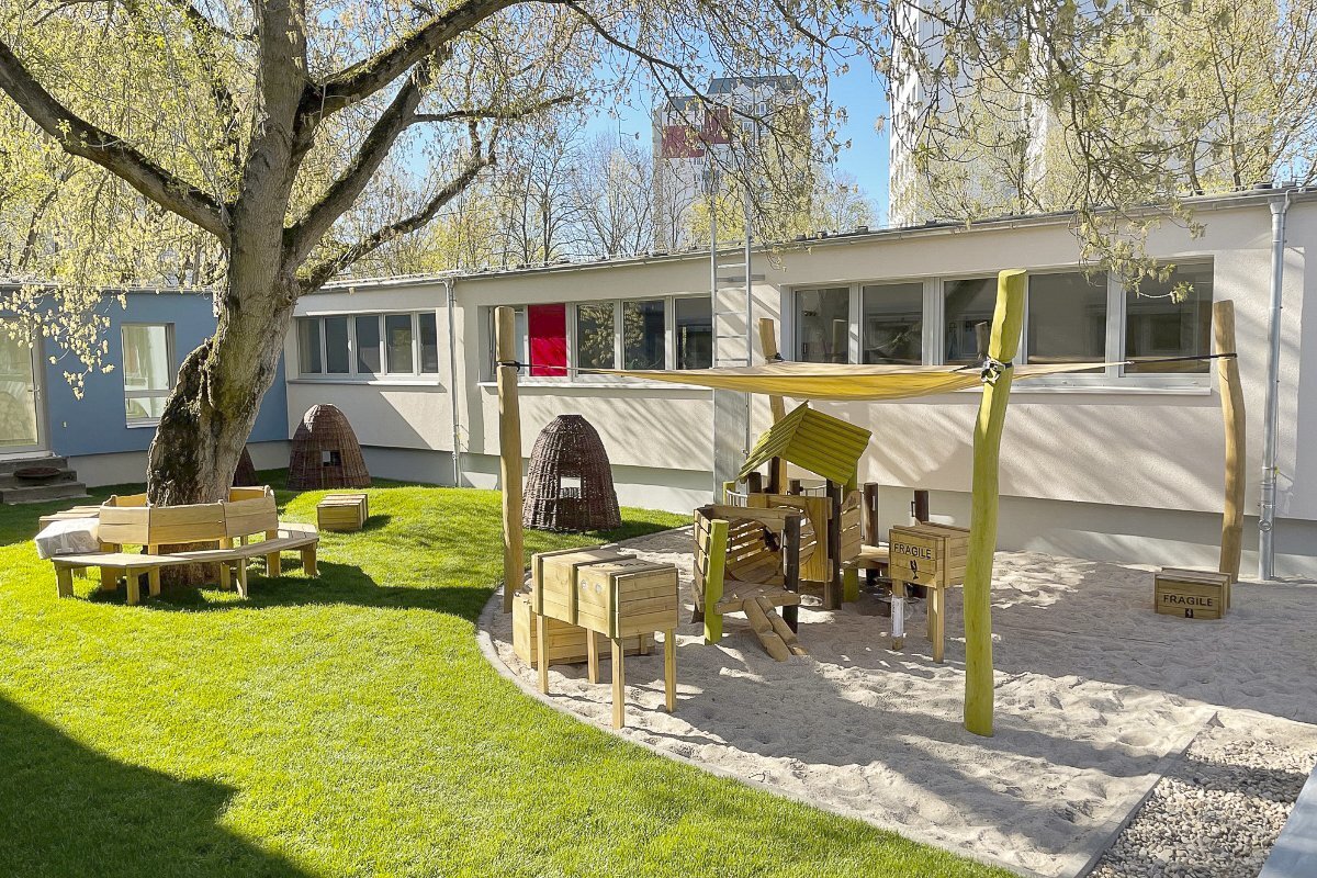 Playground equipment for childcare facilities from eibe - wooden play equipment under a sun awning, seating arrangement around a tree.